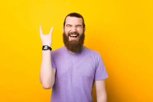 Excited man with beard in tshirt showing rock and roll gesture with fingers as if listening to favourite music