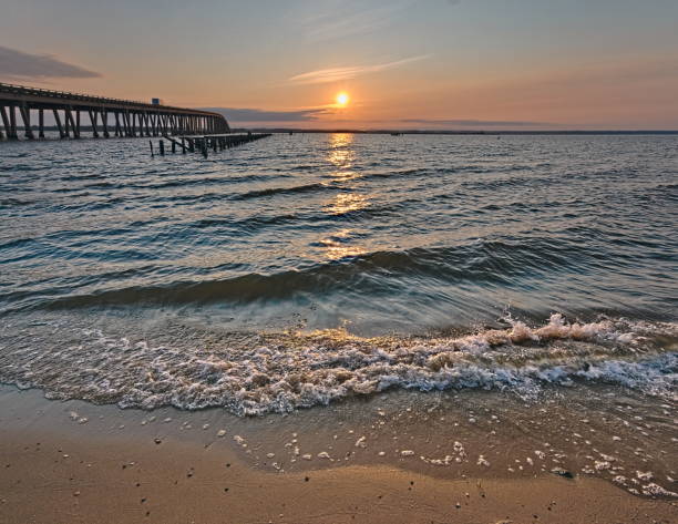 Orange Sunrise on a Rappahannock river beach.  Downing bridge and pier fading into horizon.  Waves gently washing up on the river bank.