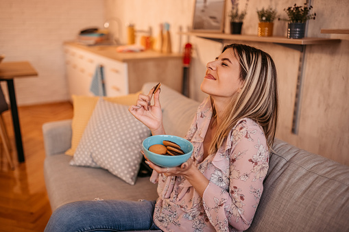 Portrait of beautiful emotional charming attractive sweet woman sitting on sofa in living room and eating cookies.