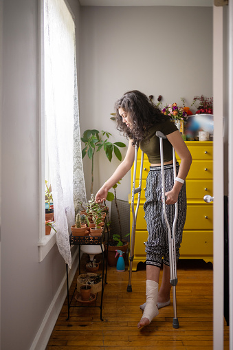 This is a photograph of a Puerto Rican Millennial woman standing up with crutches admiring her potted succulent plants by window in Chicago, USA.