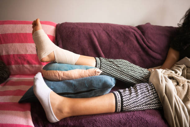 Injured Hispanic Woman Elevating Bandaged Sprained Angle on the Sofa This is a close up photograph of an injured, Millennial Puerto Rican woman resting her sprained ankle at home on a spring day in Chicago, USA. sprain stock pictures, royalty-free photos & images