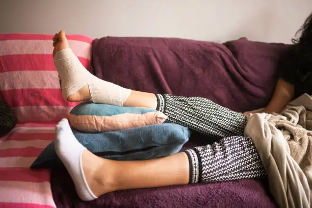 Photo of Injured Hispanic Woman Elevating Bandaged Sprained Angle on the Sofa