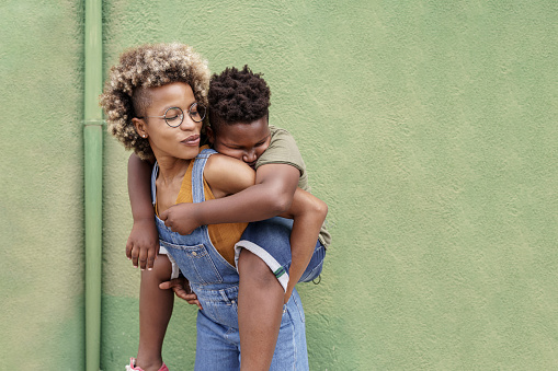 African American Young Loving Single Mother piggybacking sleepy cute little black Son in front of green wall