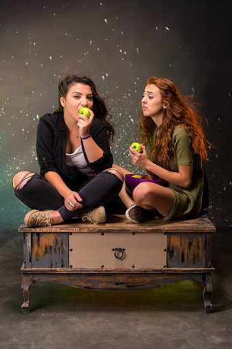 Two young girls at a fun photo shoot in the studio.
Girls with funny facial expressions making them laugh with their poses on the coffee table. Funny girls eating green apple.