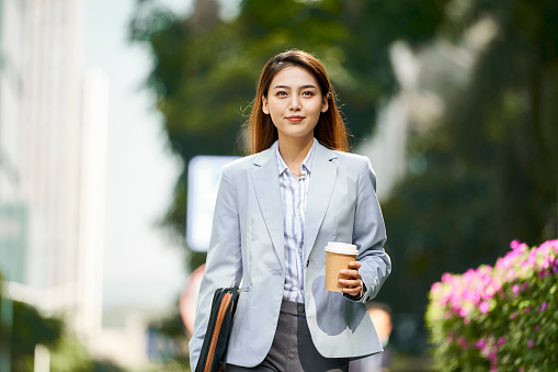 successful young asian female corporate executive walking on street holding a cup of coffee looking at camera