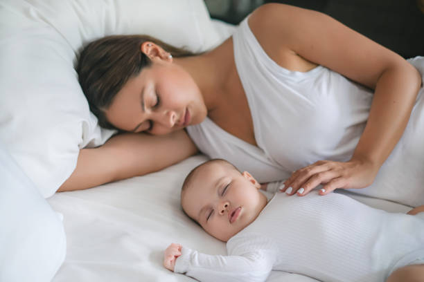 Close-up shot of beautiful new mother sleeping next to her newborn baby girl on a white bed.