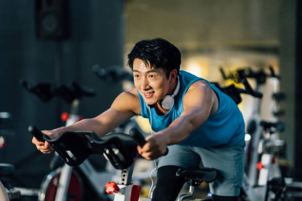 азиатский юноша катается на велотренажере в тренажерном зале. концепция здоровья и фитнеса. - exercising sport gym spinning стоковые фото и изображения
