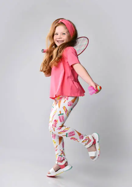 Photo of Active red-haired kid girl in pink t-shirt and colorful pants is walking, jumping with badminton racquet on shoulder