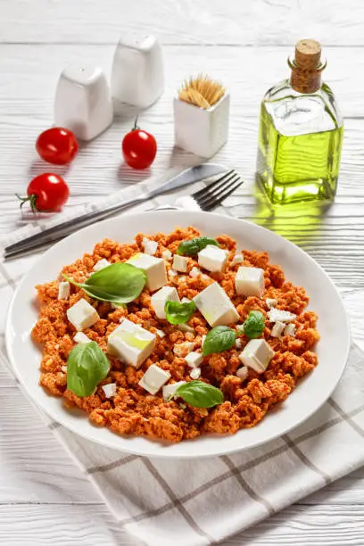 Strapatsada, eggs scrambled with tomatoes and feta cheese on a white plate with fresh basil leaves, vertical view from above, greek recipe, close-up