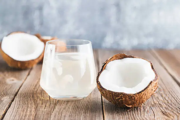 Photo of Coconut water in a glass close-up on a wooden table and a half of a fresh coconut lies nearby. Organic Coconut Detox Juice