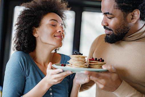 African American woman smelling fresh pancakes with her boyfriend during morning time in dining room.