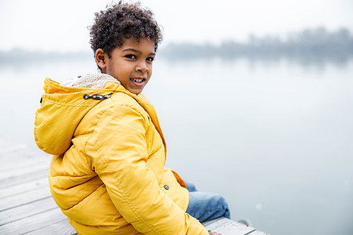 Happy black boy relaxing on a jetty in winter day and looking at camera. Copy space.