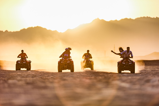 Large group of happy friends having fun while driving quads in summer at the desert at sunset. Copy space.