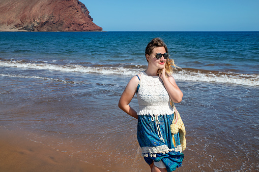 Dreamy holidays portrait of a young millennial woman wearing a blue and white dress and sunglasses while holding her sandals in one hand and walking on Tejita beach in Tenerife, Canary Islands, Spain.