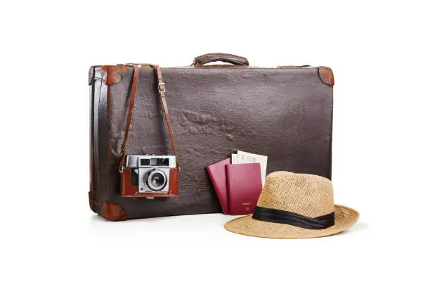Photo of Old fashioned vintage brown leather suitcase with a film camera, a straw hat and two passports with airplane tickets on white