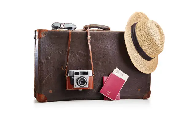 Photo of Old fashioned vintage brown leather suitcase with a film camera, a straw hat and two passports with airplane tickets on white