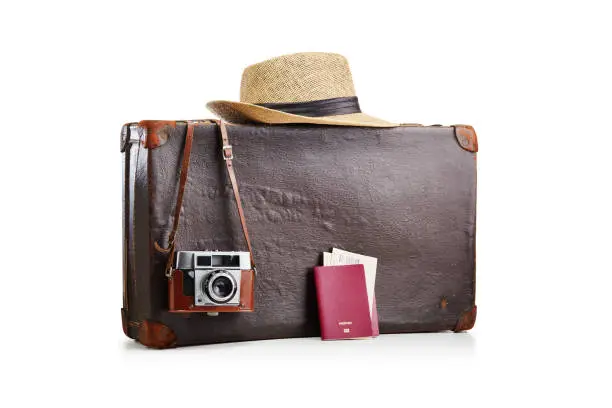 Photo of Old fashioned vintage brown leather suitcase with a film camera, a straw hat and two passports with airplane tickets on white