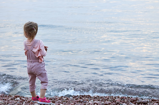 Cute little blonde girl in a pink jumpsuit stands alone on the beach and looks into the distance.