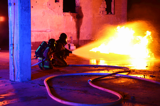 Firemen spraying water onto a big fire. Two firefighters kneeling near the flames in a protective suits and helmets.