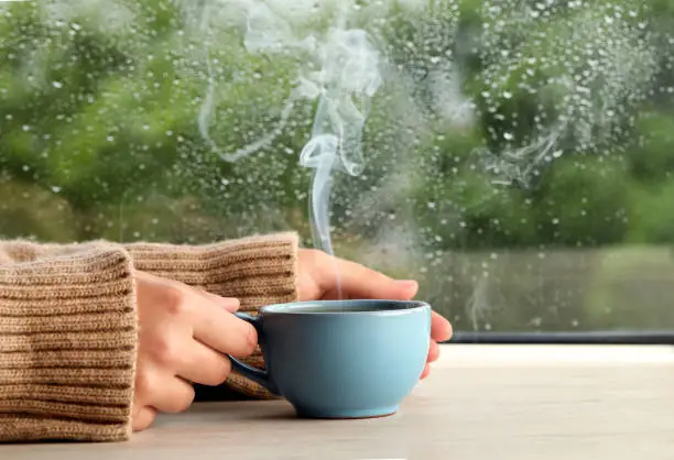Photo of Woman with cup of hot drink at wooden table near window on rainy day, closeup