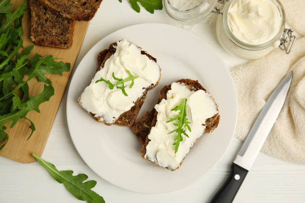 istock Bread with cream cheese and arugula on white wooden table, flat lay 1328638586