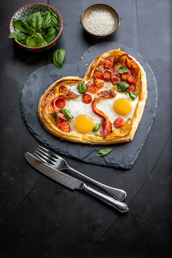 Heart shaped pizza with 2 fried eggs on top on a table surrounded by fresh ingredients.