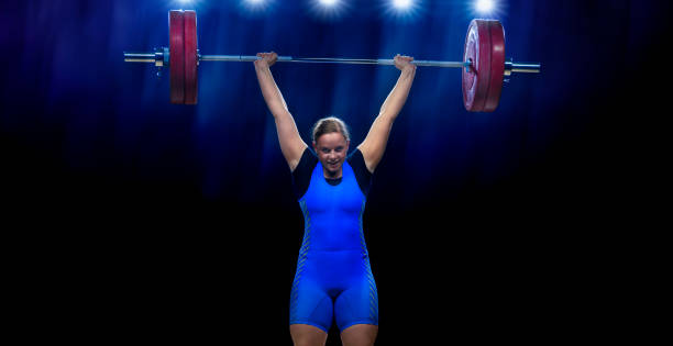 Young female weightlifter lifting barbell Portrait of smiling young female weightlifter lifting barbell above her head while performing clean and jerk lift. clean and jerk stock pictures, royalty-free photos & images