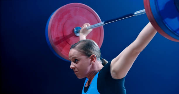 Close-up young female weightlifter lifting barbell Side view of young female weightlifter lifting barbell above her head while performing clean and jerk lift. clean and jerk stock pictures, royalty-free photos & images