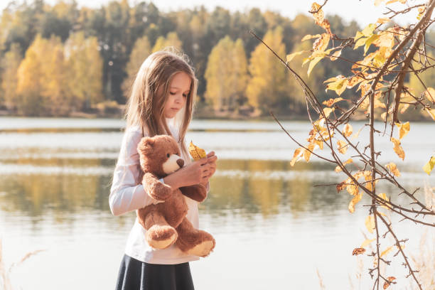 Pretty little girl in school uniform with cute teddy bear holds dry leaf on riverbank in sunny forest on autumn day side view