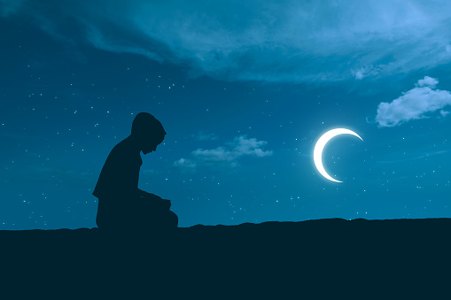 Silhouette of Muslim woman in a veil sitting and praying with crescent moon background
