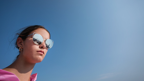 Side portrait of young lady with disheveled and uncombed hair wearing sunglasses on blue sky background with copy space outdoor