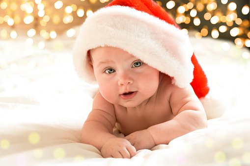 Beautiful little child is celebrating Christmas. New Year's holiday. A child in a Christmas costume. happy Newborn baby in Santa hat over holidays lights background. Horizontal banner with copy space.