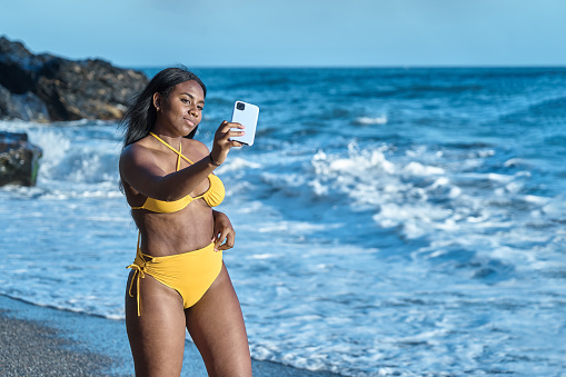 young african american woman takes a selfie on the beach shore in yellow bikini