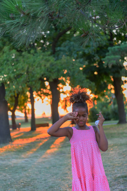 Cute Little Girl is Rubbing her Eyes in Public Park. Little African-American Curly Haired Girl with Eyes Problems is Feeling Displeased and Rubbing her Eyes During a Walk in City Park During a Summer Day. human eye scratching allergy rubbing stock pictures, royalty-free photos & images