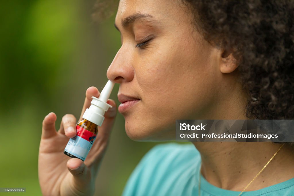 Young African-American Woman with Asthma is Using Inhaler in a Park. An African Woman in Yellow Dress is Walking in Public Park and Using an Asthma Inhaler Due to the Problems with Breathing. Spray Stock Photo