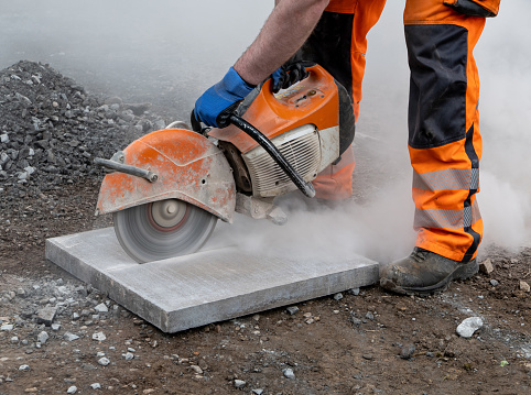 A construction worker in bright orange reflective work clothes and work boots, bending over as he uses a handheld petrol concrete grinder to cut a square paving stone to size. The heavy industrial work tool has a sharp and fast rotating circular saw, with a safety cover to protect the worker. The grinder is sending out a cloud of cement dust as it cuts through the heavy concrete stone.