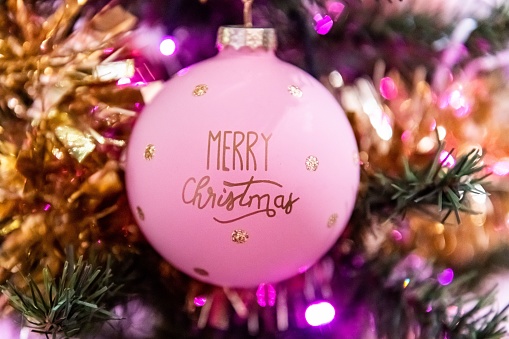 Close-up of pink Christmas ball ornament with text - Merry Christmas hanging on Christmas tree. Christmas ornament, gold chain, pink Christmas lights.