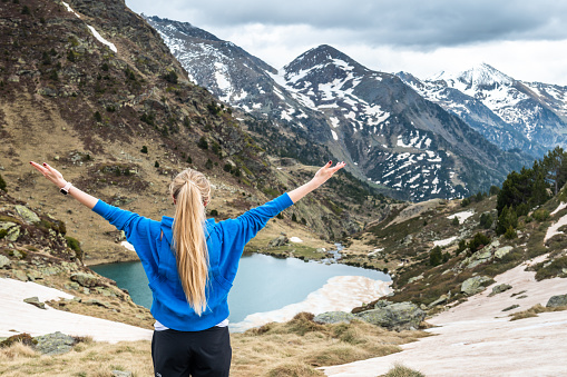 Rear view of young beautiful woman standing with arms outstretched and looking at beautiful Tristaina lake view in Pyrenees mountains, Andorra.