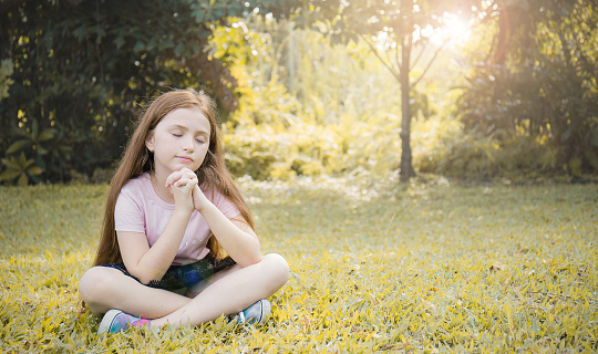 Portrait of little young caucasian girl praying in the spring park, Green grass background. Religion believe god hope concept