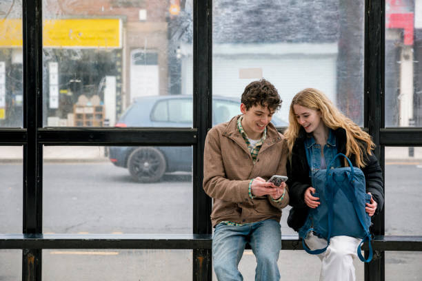 Passing the Time A front-view shot of two teenage friends sitting at a bus shelter, waiting for the bus in Wallsend, North East England. They are looking at a mobile phone together and talking. bus shelter stock pictures, royalty-free photos & images