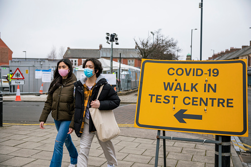A front-view shot of a COVID-19 walk-in test centre information sign in Wallsend, North East England. It is directing people where to go for a COVID-19 test. A teenage boy and girl wearing protective face masks can be seen walking along the path nearby.