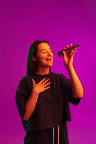 Photo of Vertical portrait of pretty young caucasian girl singing emotionally into microphone over pink background in neon lights