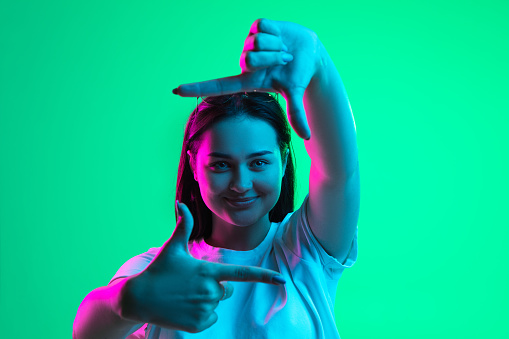 Retrato en primer plano de una joven caucásica bastante sonriente que muestra el gesto del marco aislado sobre fondo verde en luz de neón. photo