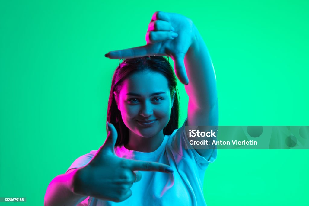 Retrato en primer plano de una joven caucásica bastante sonriente que muestra el gesto del marco aislado sobre fondo verde en luz de neón. - Foto de stock de Personas libre de derechos