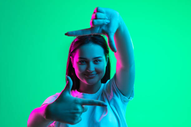 Close-up portrait of young pretty smiling caucasian girl showing frame gesture isolated on green background in neon light. Pleasant emotions. One young beautiful girl demonstrating frame gesture isolated over light green background in neon light. Close-up image Gesturing stock pictures, royalty-free photos & images