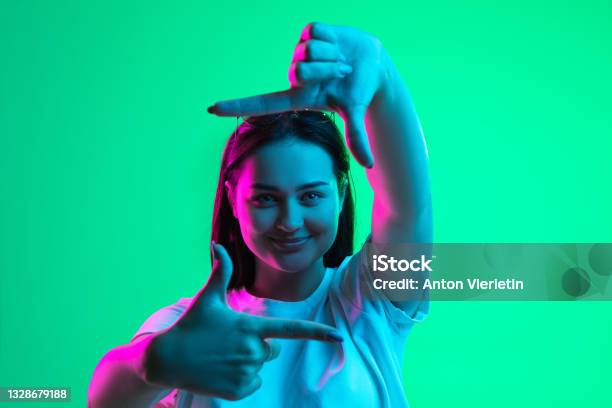 Closeup Portrait Of Young Pretty Smiling Caucasian Girl Showing Frame Gesture Isolated On Green Background In Neon Light Stock Photo - Download Image Now