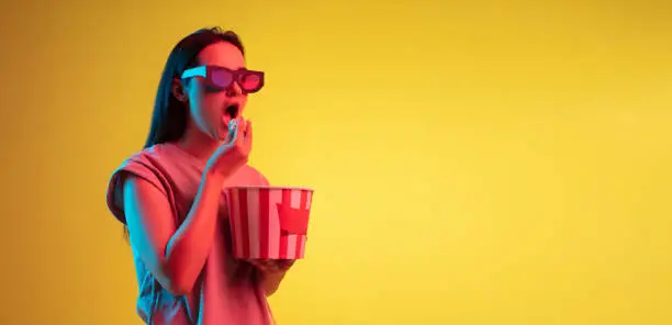 Photo of Watching cinema. Young caucasian girl in 3d glasses eating popcorn isolated on yellow background in neon light. Flyer