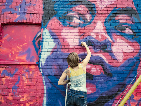 Young woman painting mural using spray paints. She is dressed in casual work clothes. Exterior of urban street, back alley with old brick wall of the house.