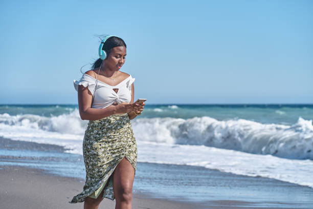 young african american woman listening to music by the shore of the beach stock photo