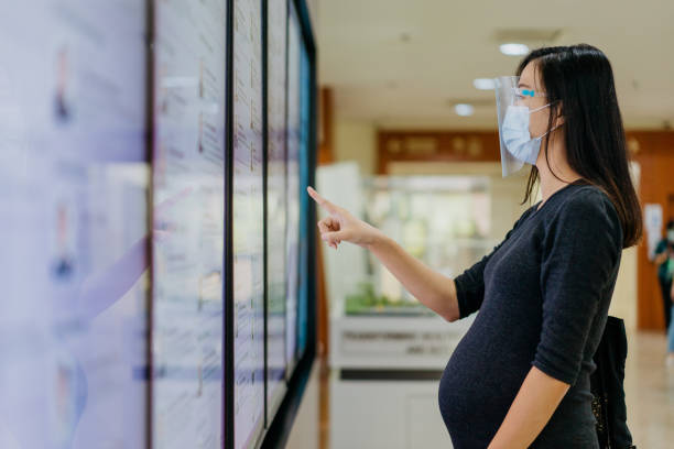 donna incinta con maschera facciale e scudo facciale con chiosco touch screen nel centro commerciale - human pregnancy clothing shopping retail foto e immagini stock
